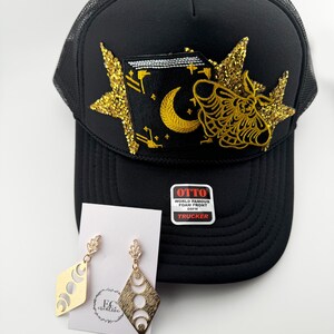 May include: Black trucker hat with a gold glitter star and a gold moth and moon design. The hat has a label that says "Otto World Famous Foam Front OSFM Trucker".  The image also shows a pair of gold earrings with a crescent moon design.