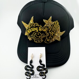 May include: Black trucker hat with a gold glitter patch that says "Witching Hour" and a gold glitter hand with stars.  A pair of black snake earrings with gold glitter accents and a crescent moon design.