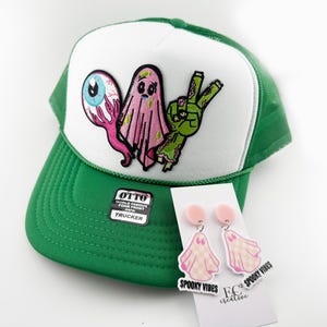 May include: Green and white trucker hat with a patch featuring a cartoon eye, ghost, and peace sign. Includes a pair of pink ghost earrings with the text "Spooky Vibes".