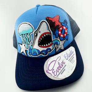 May include: A blue and navy blue trucker hat with a shark, jellyfish, and anchor design. The hat has a concealed ponytail opening and the text "Evolve Eluded" on the tag.
