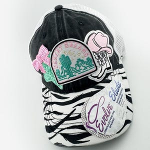 May include: Black and white zebra print baseball cap with a black denim crown and a white mesh back. The cap has several patches including a pink cowboy boot, a pink bow, and a green cactus with the text "Desert Dreams". The cap has a label that says "Evolve Eluded" with the text "Concealed Ponytail Opening".