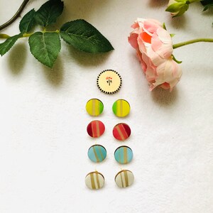 May include: A selection of handmade wooden stud earrings in various colours and striped designs. The earrings include yellow, green, red, blue, and cream colours. A small, round wooden pin with a floral design is also present.