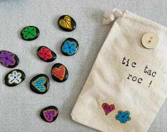 Può includere: Un set di sassi dipinti con motivi floreali e a cuore colorati, vicino a un sacchetto di tela. La borsa è stampata con "tic tac roc!" e ha un bottone di legno con un fiore. Blocchi di legno giocattolo sullo sfondo.