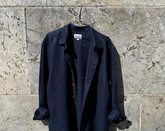 May include: A navy blue button-down shirt with long sleeves, hanging on a wire hanger against a textured stone wall. The shirt has a relaxed fit, rolled-up sleeves, and the brand name "toel" is visible on the label.