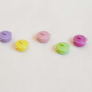 May include: Five miniature pastel-coloured pots with white handles. The pots are arranged in a row from left to right: purple, yellow, pink, green, and purple.
