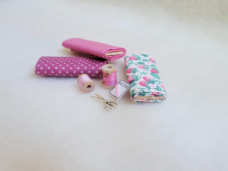 May include: A set of pink fabric scraps, two spools of pink thread, a pair of silver scissors, and a small card that says "Buttons".