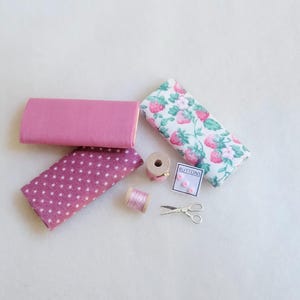 May include: A collection of pink fabric scraps, a spool of pink thread, a small box of buttons labeled "Buttons", and a pair of silver scissors.