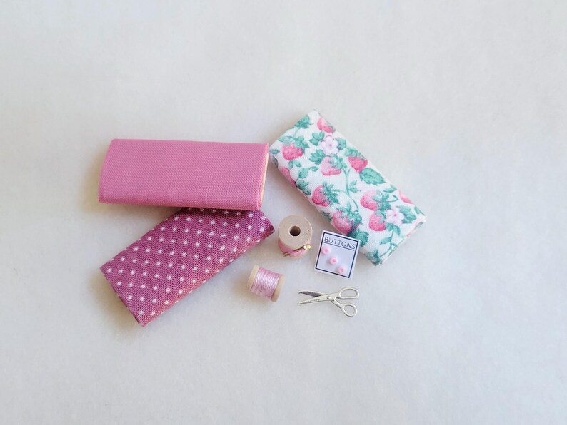 May include: A collection of pink fabric scraps, a spool of pink thread, a small box of buttons labeled "Buttons", and a pair of silver scissors.