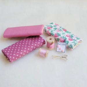 May include: A set of pink fabric scraps, two spools of pink thread, a small spool of thread with a needle, a card with two pink buttons and the word "BUTTONS" printed on it, and a pair of silver scissors.