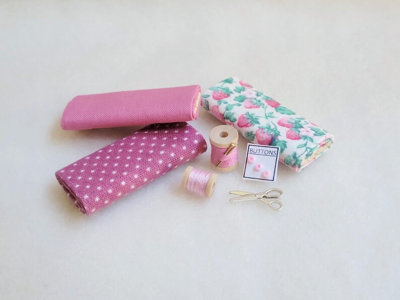 May include: A set of pink fabric scraps, two spools of pink thread, a small spool of thread with a needle, a card with two pink buttons and the word "BUTTONS" printed on it, and a pair of silver scissors.