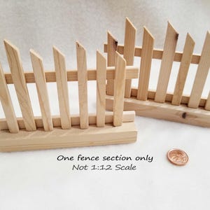 May include: Two sections of a miniature wooden fence, each with a base. The fence is made of thin, light brown wooden slats. The fence is approximately 10 cm tall. One fence section is leaning against the other. The text "One fence section only Not 1:12 Scale" is printed below the fence.