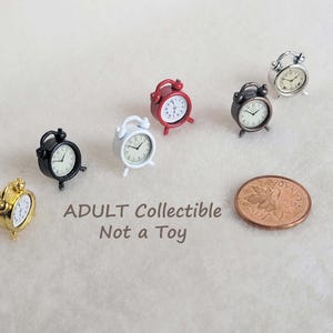 Puede incluir: Una colección de seis despertadores en miniatura en oro, negro, blanco, rojo, gris oscuro y plata, cada uno con esfera blanca y manecillas negras. Se incluye una moneda de un centavo canadiense de cobre para escala. El texto ADULT Collectible Not a Toy es visible.