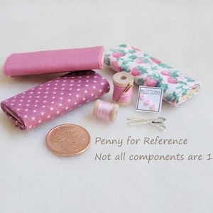May include: A collection of miniature sewing supplies, including pink fabric, spools of thread, buttons, and a pair of scissors. A penny is included for scale.