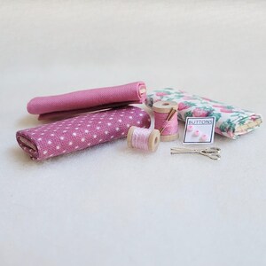 May include: A set of pink and white fabric scraps, two spools of pink thread, a small pack of pink buttons labeled "BUTTONS", and a pair of silver scissors.