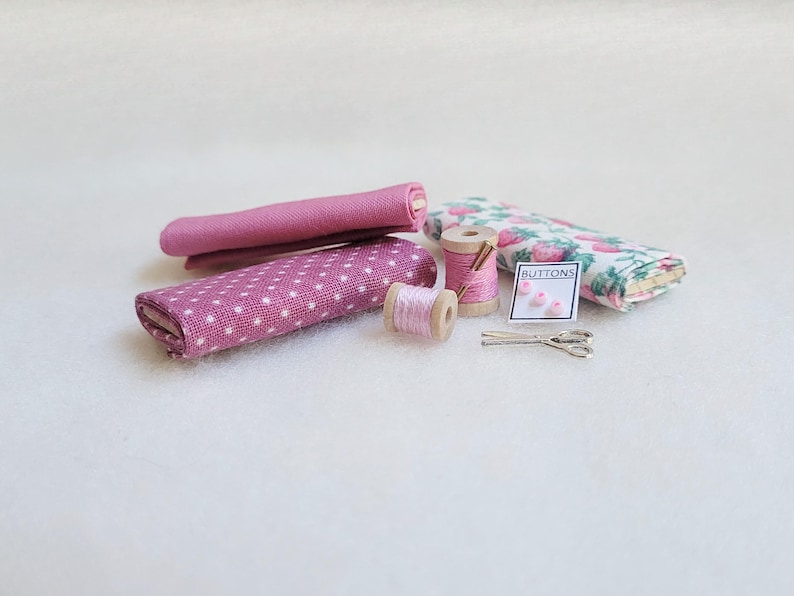 May include: A set of pink and white fabric scraps, two spools of pink thread, a small pack of pink buttons labeled "BUTTONS", and a pair of silver scissors.