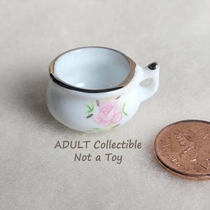 May include: A miniature white ceramic chamber pot with a silver-toned rim and a small handle. The exterior features a painted pink rose with green leaves. Next to the item is a Canadian one-cent coin for scale. Text reads: ADULT Collectible Not a Toy.