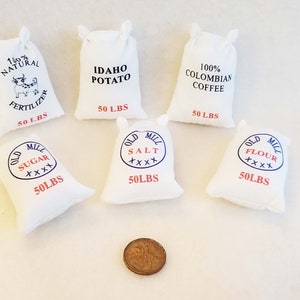 May include: Six white fabric sacks with printed labels. The sacks are labelled with various products, including "Idaho Potato", "100% Colombian Coffee", "Old Mill Sugar", "Old Mill Salt", "Old Mill Flour", and "100% Natural Fertilizer". Each sack is labelled with "50 LBS".