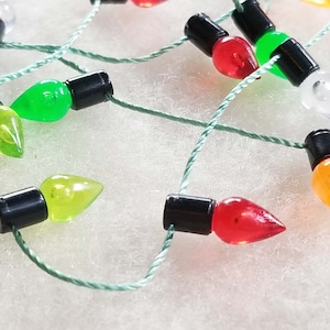 May include: Close-up of a string of Christmas lights with various coloured bulbs, including green, red, yellow, and clear. Each bulb has a black base and is strung on a green wire. The lights are arranged against a white background.
