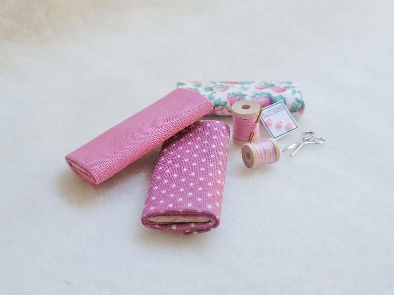 May include: A set of pink fabric scraps, two spools of pink thread, a small package of buttons labeled "Buttons", and a pair of silver scissors.