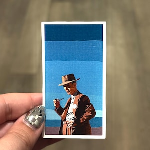May include: A rectangular sticker featuring a stylized illustration of a person in a brown suit and hat, holding a pipe. The background has blue and purple horizontal stripes. The sticker is held by a hand with a silver and black checkered nail.