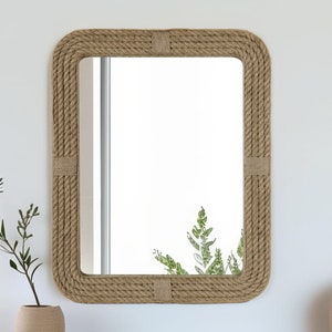 May include: A rectangular mirror with a thick rope frame. The frame is made of natural colored rope and has a woven texture. The mirror is reflecting a plant.