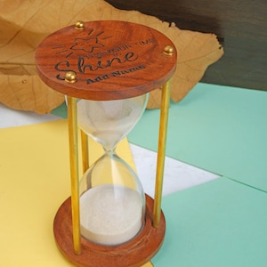 May include: A wooden hourglass with gold accents and a motivational quote that reads "It's your time to shine" with a space to add a name. The hourglass is sitting on a yellow and blue surface.
