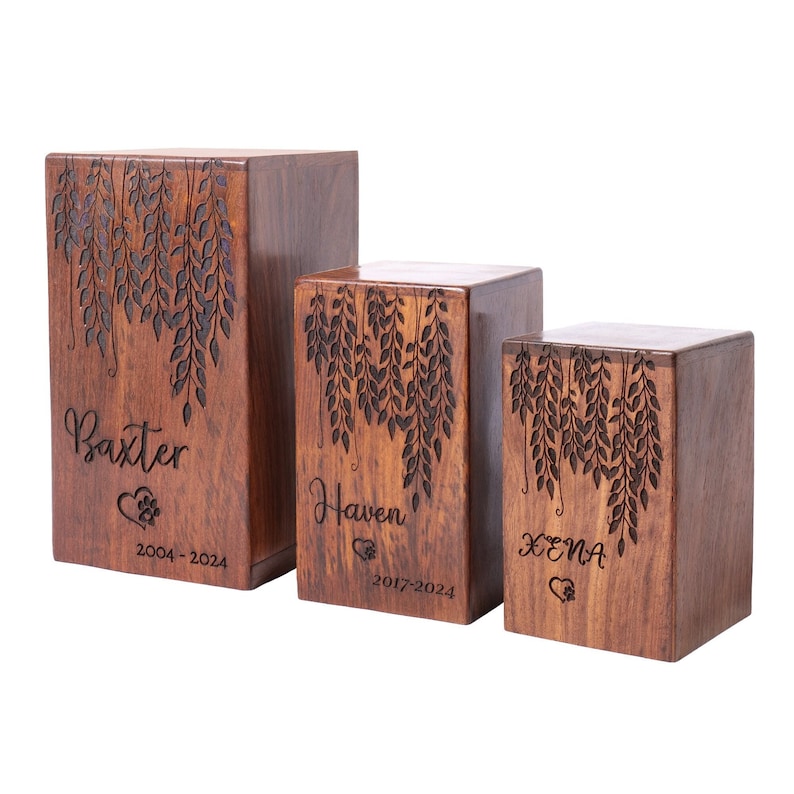 May include: Three wooden pet urns with engraved names, dates, and a paw print. The urns are decorated with a design of hanging leaves. The urns are in different sizes, with the largest urn in the back and the smallest urn in the front. The urns are all made of a dark wood.