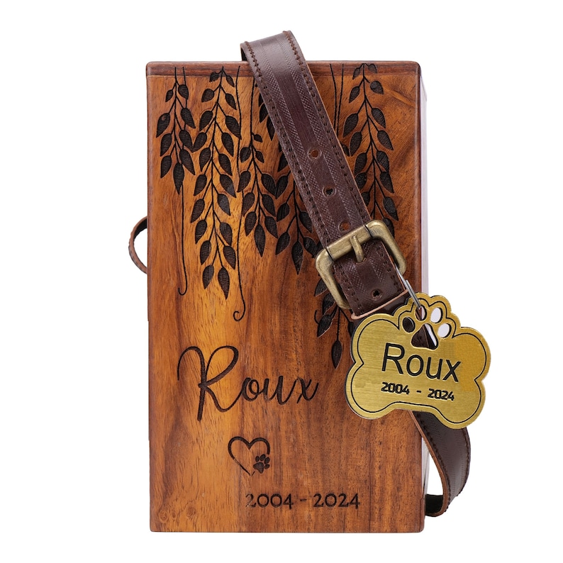 May include: A wooden pet urn with a brown leather strap and a gold metal tag that says "Roux 2004 - 2024". The urn has a carved design of leaves and the name "Roux" is engraved on the front.