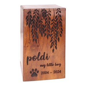 May include: A wooden pet urn with a carved design of leaves and branches. The urn is engraved with the name "poldi", "my little boy", and the dates "2004 - 2024".