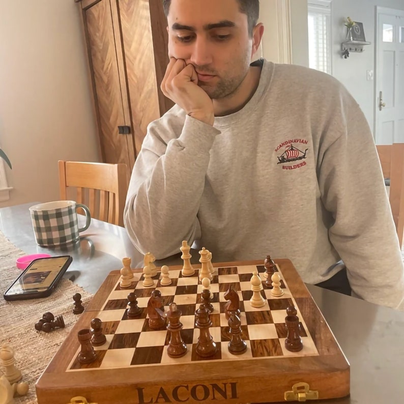 May include: A wooden chess set with a chessboard and chess pieces is displayed on a table. The chessboard has a light and dark wood pattern. The chess pieces are brown and white. The word "LACONI" is printed on the chess set.