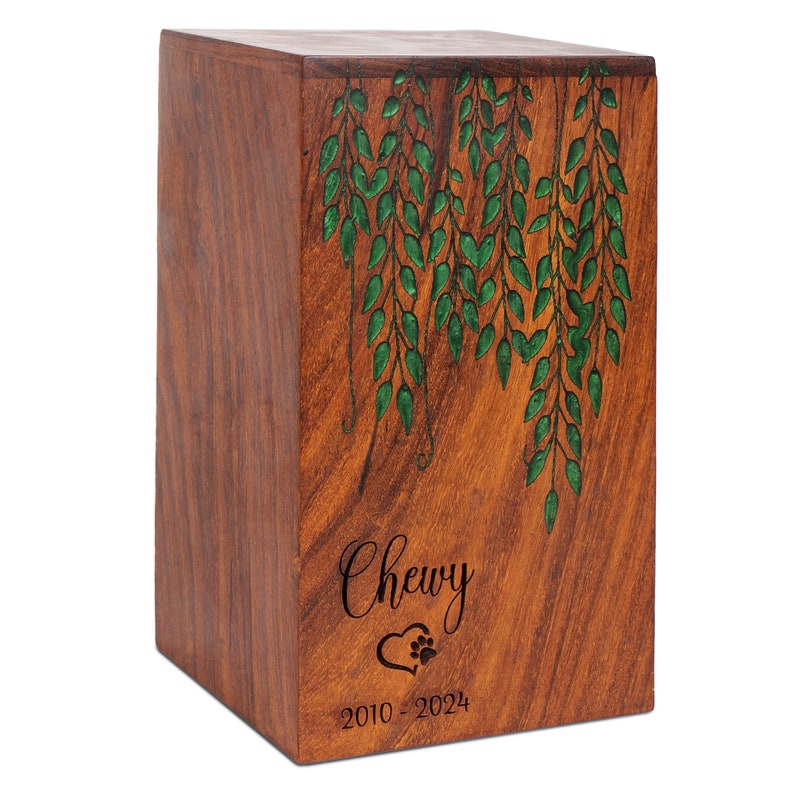 May include: A wooden pet urn with a green leafy design and the name "Chewy" engraved on the front. The urn also includes the dates "2010 - 2024" and a heart with a paw print.
