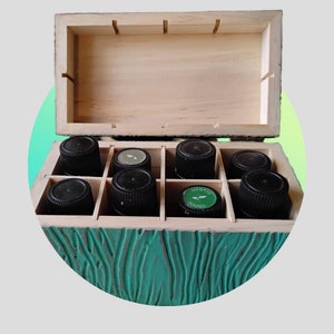 Peut inclure: Une boîte en bois avec un couvercle vert sarcelle et un motif en bois. La boîte est ouverte et contient huit petits flacons en verre avec des bouchons noirs. Un flacon porte une étiquette verte sur laquelle est inscrit "MENTHE POIVREE".