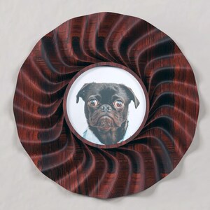May include: A circular award ribbon featuring a dark brown and black pleated fabric rosette. At the center is a round white insert displaying a detailed illustration of a black pug dog with large eyes, wearing a light blue collared shirt.
