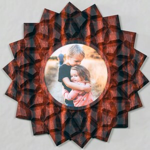 May include: A circular wall frame featuring a dark red and black buffalo plaid pattern with a starburst or sunburst design. The frame has a central circular opening displaying a photograph of two children embracing in an outdoor setting.