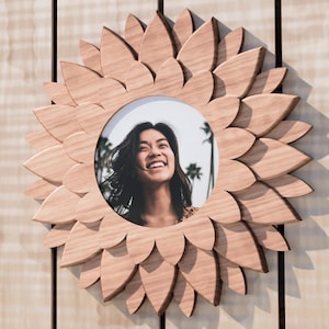 May include: A wooden picture frame shaped like a sunflower with a photo of a woman smiling inside. The frame is a light brown color and has a natural wood grain.