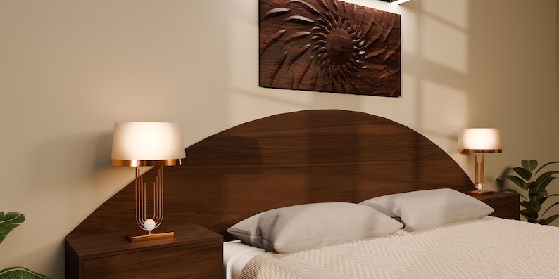 May include: A dark wood arched headboard with matching bedside tables, each featuring a gold-toned lamp with a white cylindrical shade. A textured white bedspread with two pillows sits in front of the headboard, beneath a dark wood wall art piece.