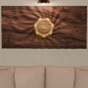 May include: A rectangular wall art piece featuring a dark brown wood grain background with a central, circular gold-colored emblem. The emblem displays a symmetrical, radiating floral or sunburst pattern with intricate, curved, petal-like details.