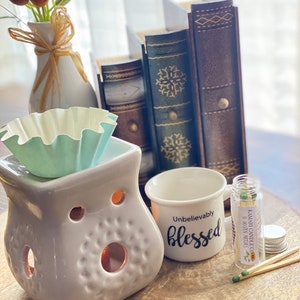 May include: A white ceramic oil diffuser with a teal wax melt warmer, a white mug with the text "Unbelievably Blessed", a small glass jar with matches, and a stack of leather-bound books.