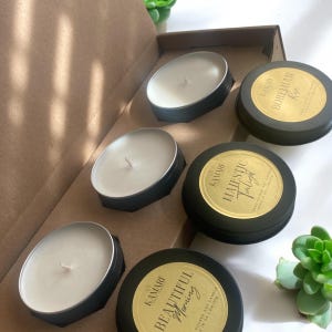 May include: A set of four soy candles in black tins with gold labels. The labels have the names "Beautiful Morning", "Majestic Twilight", "Bohemian Rose", and "Rose".