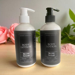 May include: Two white body lotion bottles with black labels. The labels read "BODY LOTION" and "BLUSH SUGAR". One bottle has a white pump, the other a black pump. The background includes pink roses and pink granules.