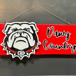 May include: A rectangular red wooden sign featuring a white and black bulldog head illustration with a spiked collar and the letter G, alongside the black text Dawg Country. The sign is suspended by a thin silver wire for wall mounting.
