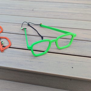 May include: Two pairs of glasses, one orange and one green, are lying on a wooden surface. Both pairs have a flexible frame and a black cord.
