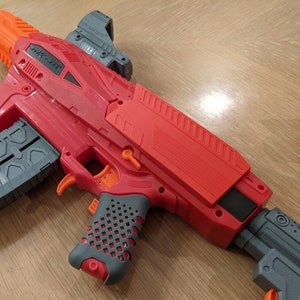 Op de afbeelding: Een rood en grijs speelgoedpistool met een zwart magazijn waarop "DZP - 15" staat. Het pistool heeft een richtkijker bovenop en een getextureerde grip.