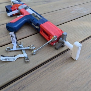 May include: A red and blue toy gun with a white handle and a silver wrench. The gun is lying on a wooden surface.