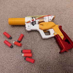 Puede incluir: Una pistola de juguete impresa en 3D de color blanco, amarillo y rojo con un mango rojo y un cañón amarillo. La pistola está rodeada de dardos de espuma rojos y blancos.