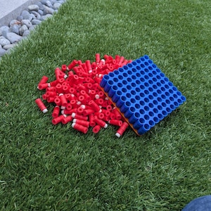 May include: A pile of red toy darts with white tips next to a blue dart holder on green artificial turf. The dart holder is rectangular with many circular holes.