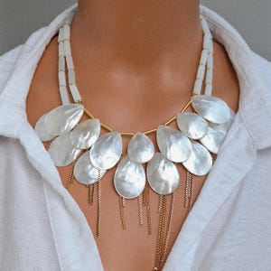 May include: A statement necklace featuring large, iridescent, teardrop-shaped pendants. The pendants are arranged in a cascading design, suspended from a gold-tone metal frame. The necklace includes delicate gold chain fringe and a white beaded strand.