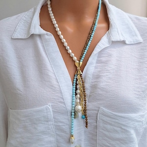 Peut inclure: Un collier à plusieurs rangs avec des perles blanches, des perles bleu clair et des perles marron. Le collier a un fermoir doré et un design en forme de gland. Le collier est porté sur une chemise blanche.