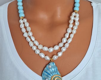 May include: A layered necklace featuring light blue beads, white pearls, and a blue seashell pendant with gold accents. The necklace is displayed on a white background.