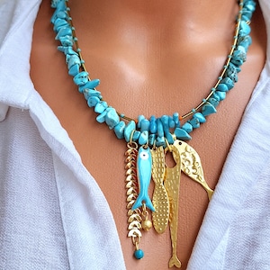 Pode incluir: Um colar de pedras turquesas com pingentes dourados de peixes e folhas. O colar apresenta uma dupla corrente de lascas de turquesa e detalhes metálicos dourados. Os pingentes de peixe são dourados e azul claro.